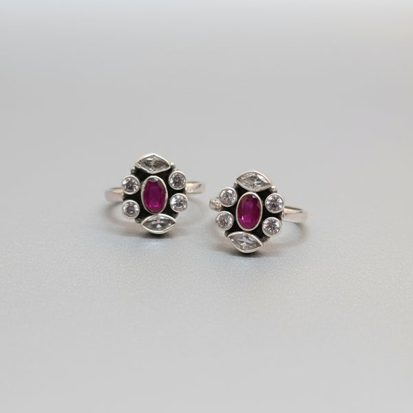 A pair of 92.5 silver toe rings adorned with red stones and small white diamonds displayed on a green leaf.