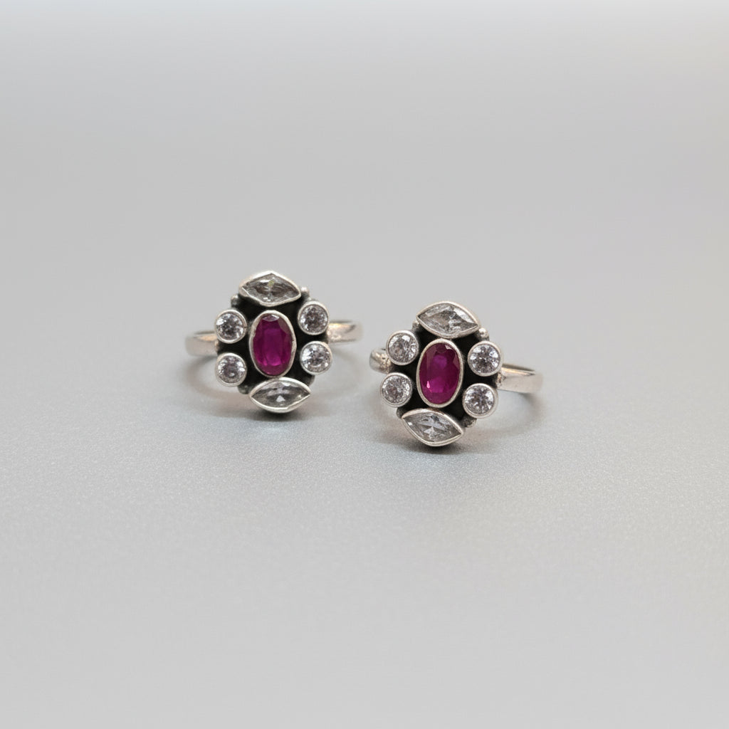 A pair of 92.5 silver toe rings adorned with red stones and small white diamonds displayed on a green leaf.