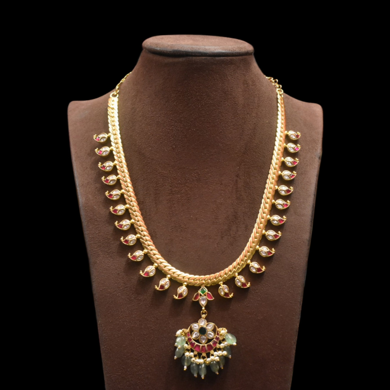 Gold necklace with green and red accents on a brown mannequin against a black background