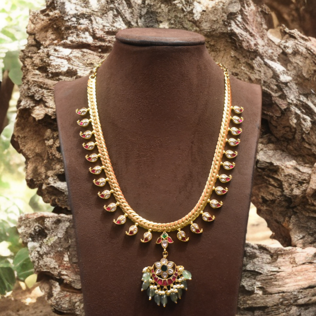 Gold necklace with green and red accents on a brown mannequin against a natural background
