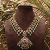 Ornate necklace with green and red stones on a brown mannequin against a leafy background