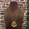 Decorative necklace with green and gold elements on a brown mannequin against a brick wall.