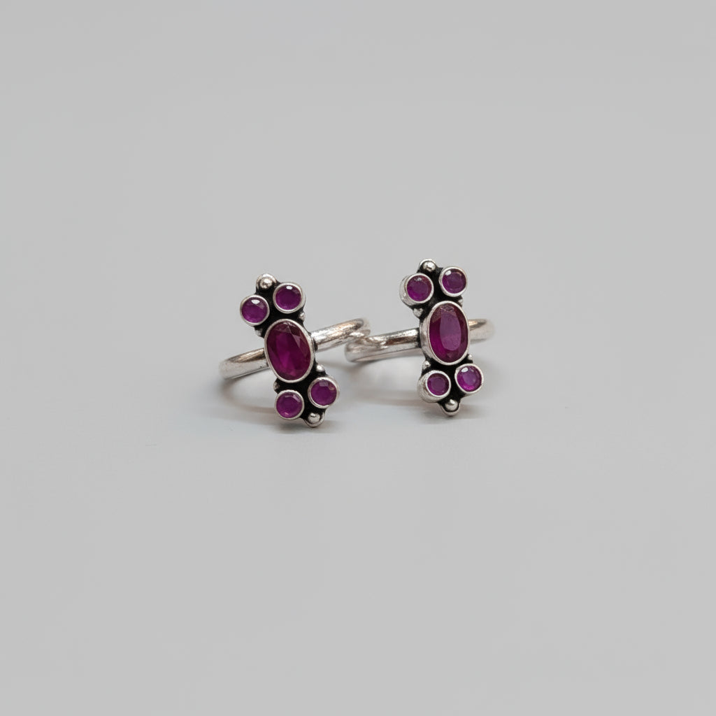 A pair of 92.5 silver ruby and emerald adorned bichiya toe rings placed on a red leaf background.